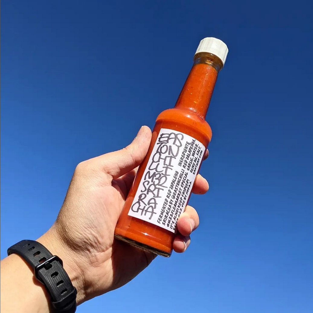 Hand holding a bottle of Jalapeno Sriracha sauce aloft against a blue sky.